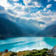 Turquoise alpine lake surrounded by snow-capped mountains and forested valleys under blue sky with white clouds